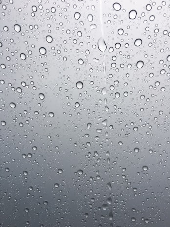 Droplets of water scattered across a smooth glass surface, with varying sizes and clear reflections. The background appears slightly blurred and gray, emphasizing the clarity and texture of the water droplets.