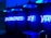 A series of blue neon lights arranged against a perforated wall, spelling the words 'POWERED BY YOU'. Industrial-style hanging lamps are visible at the top, emitting a blue glow that complements the neon text. Below, countertop appliances like a microwave are partially visible.