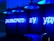 A series of blue neon lights arranged against a perforated wall, spelling the words 'POWERED BY YOU'. Industrial-style hanging lamps are visible at the top, emitting a blue glow that complements the neon text. Below, countertop appliances like a microwave are partially visible.