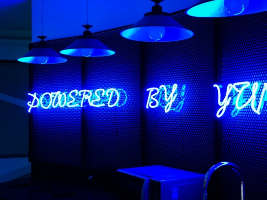 A series of blue neon lights arranged against a perforated wall, spelling the words 'POWERED BY YOU'. Industrial-style hanging lamps are visible at the top, emitting a blue glow that complements the neon text. Below, countertop appliances like a microwave are partially visible.