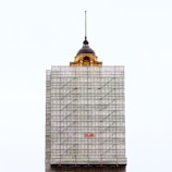 A partially completed building is covered with scaffolding. The top portion features an older architectural style with a dome and a spire, contrasting with the modern construction scaffolding. The word 'Built' is prominently displayed in red on the scaffolding.