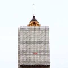 A partially completed building is covered with scaffolding. The top portion features an older architectural style with a dome and a spire, contrasting with the modern construction scaffolding. The word 'Built' is prominently displayed in red on the scaffolding.