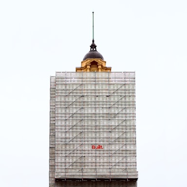 A partially completed building is covered with scaffolding. The top portion features an older architectural style with a dome and a spire, contrasting with the modern construction scaffolding. The word 'Built' is prominently displayed in red on the scaffolding.