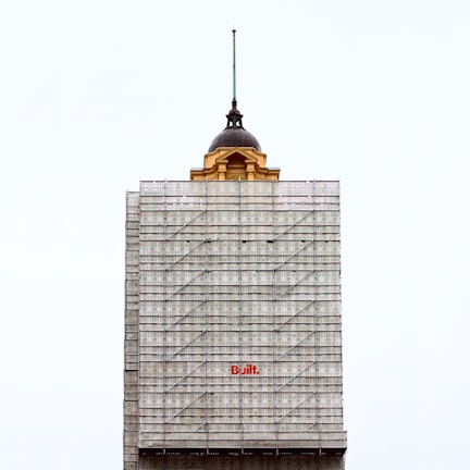 A partially completed building is covered with scaffolding. The top portion features an older architectural style with a dome and a spire, contrasting with the modern construction scaffolding. The word 'Built' is prominently displayed in red on the scaffolding.