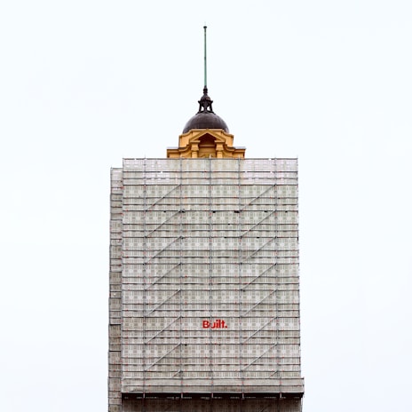 A partially completed building is covered with scaffolding. The top portion features an older architectural style with a dome and a spire, contrasting with the modern construction scaffolding. The word 'Built' is prominently displayed in red on the scaffolding.