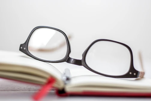 A sleek pair of black-rimmed eyeglasses resting on an open book beside a cup of coffee.