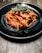 A black ceramic plate holds a serving of pasta coated in rich tomato sauce, topped with fresh basil leaves. The pasta includes chunks of tofu or a similar protein mixed with red and green bell peppers. The dish rests on a rustic wooden table.
