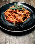 A black ceramic plate holds a serving of pasta coated in rich tomato sauce, topped with fresh basil leaves. The pasta includes chunks of tofu or a similar protein mixed with red and green bell peppers. The dish rests on a rustic wooden table.