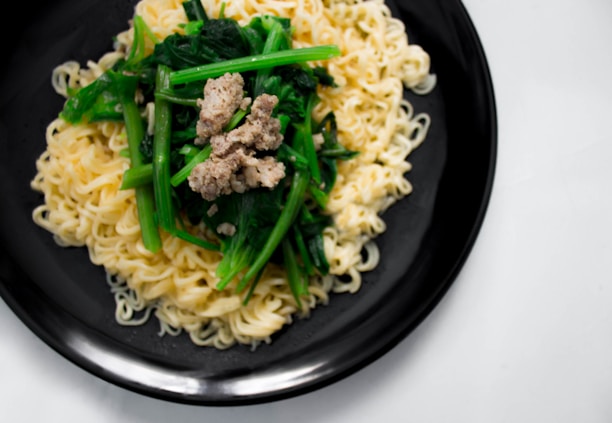 A plate contains a serving of cooked noodles topped with green vegetables and a small portion of browned ground meat. The dish is arranged neatly on a black plate, set against a light background.
