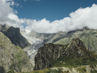 A rugged mountain landscape with steep peaks and a glacier nestled between them, beneath scattered white clouds in a vivid blue sky. The foreground features lush green grass and rocky outcrops, where a solitary hiker wearing red is visible.
