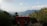 A small red building with solar panels on the roof is surrounded by lush green forests. In the background, there are layers of misty mountains under a clear blue sky with scattered clouds.