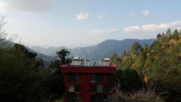 A small red building with solar panels on the roof is surrounded by lush green forests. In the background, there are layers of misty mountains under a clear blue sky with scattered clouds.