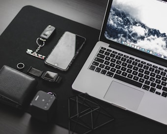 A neatly arranged collection of everyday technology items on a dark surface, including a laptop with a scenic wallpaper, a smartphone, a wallet, a key fob with a GTI keychain, a digital watch, and a small cube-shaped device.