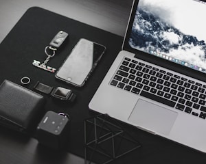 A neatly arranged collection of everyday technology items on a dark surface, including a laptop with a scenic wallpaper, a smartphone, a wallet, a key fob with a GTI keychain, a digital watch, and a small cube-shaped device.