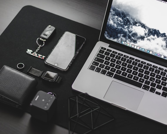 A neatly arranged collection of everyday technology items on a dark surface, including a laptop with a scenic wallpaper, a smartphone, a wallet, a key fob with a GTI keychain, a digital watch, and a small cube-shaped device.