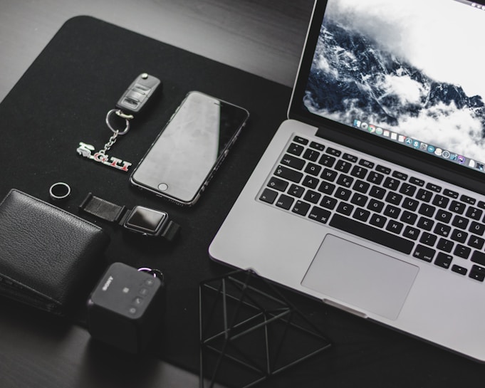 A neatly arranged collection of everyday technology items on a dark surface, including a laptop with a scenic wallpaper, a smartphone, a wallet, a key fob with a GTI keychain, a digital watch, and a small cube-shaped device.