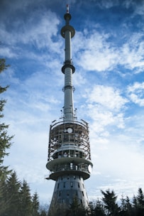 A tall communication tower stands against a backdrop of a partly cloudy sky. The structure is cylindrical with multiple satellite dishes and antennas attached. The lower section is encircled by scaffolding, indicating ongoing maintenance work. Trees surround the base, suggesting a remote or forested area.