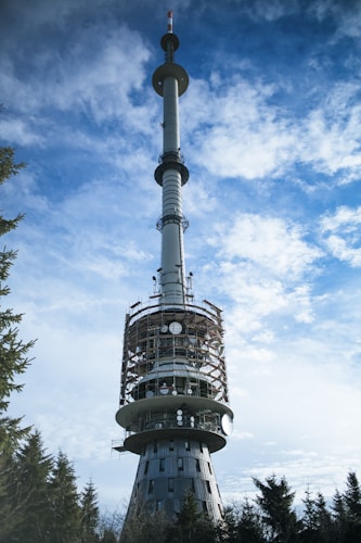 A tall communication tower stands against a backdrop of a partly cloudy sky. The structure is cylindrical with multiple satellite dishes and antennas attached. The lower section is encircled by scaffolding, indicating ongoing maintenance work. Trees surround the base, suggesting a remote or forested area.
