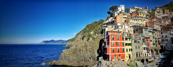 A picturesque coastal town with colorful buildings perched on the edge of a steep cliff overlooking a calm, deep blue sea. The horizon stretches with a clear blue sky meeting the sea, while rugged mountains are visible in the distance. The buildings vary in hues, including red, yellow, pink, and green, creating a vibrant and lively atmosphere.