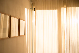 Soft beige linen curtains gently draped over a light wood window frame, bathed in natural daylight.