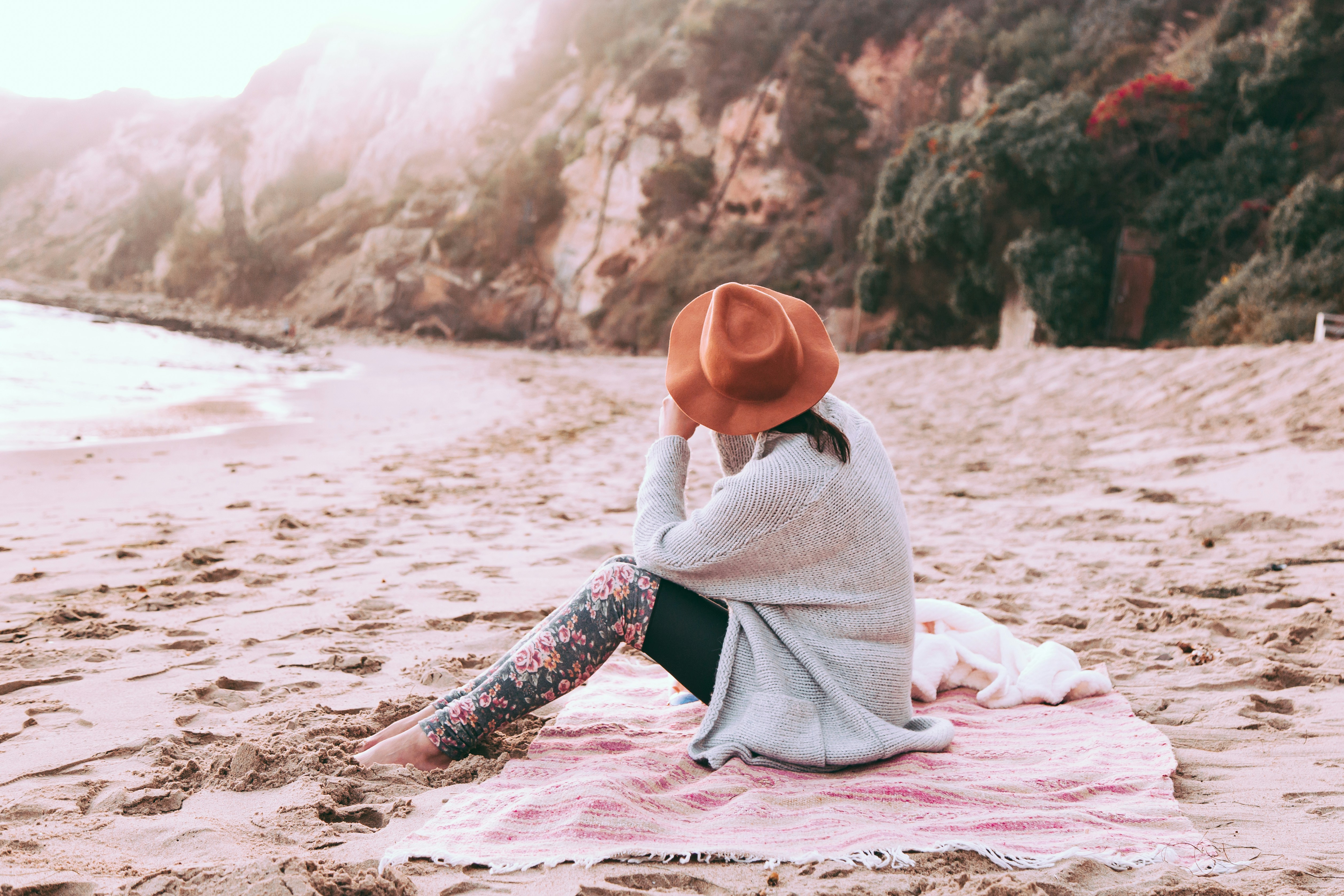 photo of person sitting on sand at seashore, Digging your feet into the sand, watching the sunset over the cliffs, and listening to the ocean is a great way to reset after a workday. This time of reflection keeps your focus and helps fuel creativity.