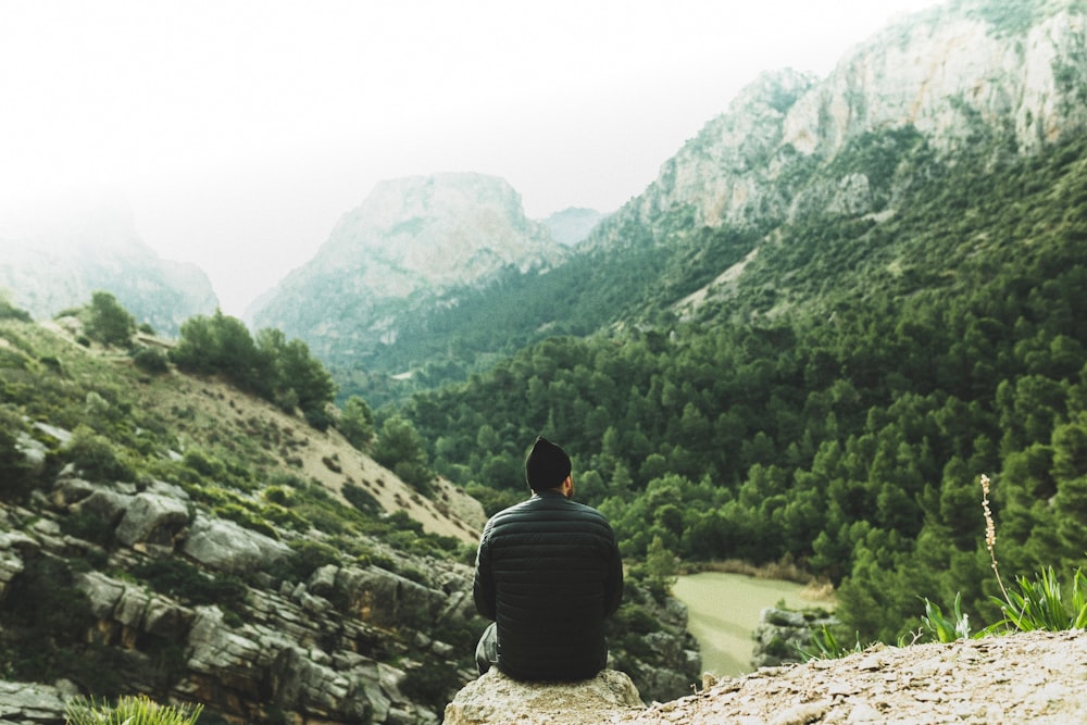 man sitting on rock looking at green trees in mountains