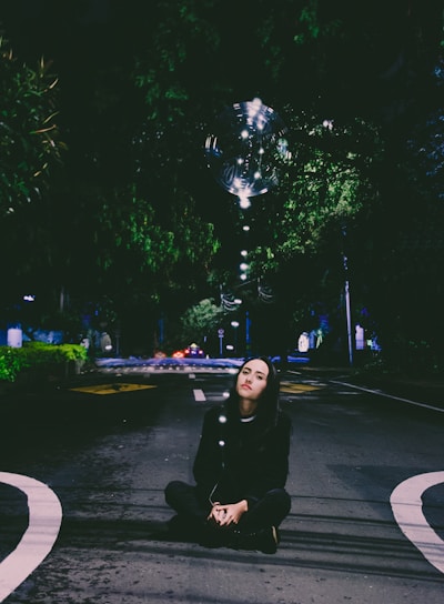 A person with long hair sits cross-legged on a dimly lit street under a canopy of trees. Above them floats a large, transparent bubble illuminated by small, white lights. The scene is serene and slightly surreal, with the dark surroundings contrasting with the bright bubble.