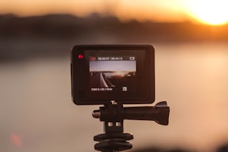 A camera mounted on a tripod is filming a sunset over a body of water. The camera's screen displays the live view of the scene with clear details of the sun setting against the horizon.