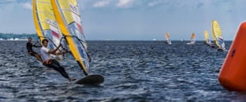 Multiple windsurfers navigate through the water, with some leaning forward and gripping their sails tightly. The background includes a distant shoreline under a partly cloudy sky, and an orange buoy is visible on the right side of the image.