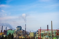 A large industrial complex with several buildings, smokestacks, and visible smoke emissions rising into the sky. The scene includes metal structures and pipework, with a clear sky overhead and trees in the foreground.