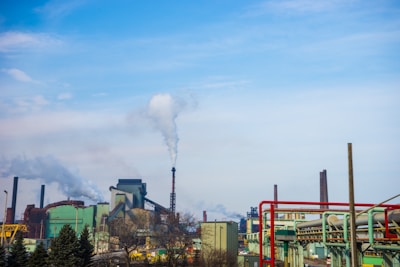 A large industrial complex with several buildings, smokestacks, and visible smoke emissions rising into the sky. The scene includes metal structures and pipework, with a clear sky overhead and trees in the foreground.