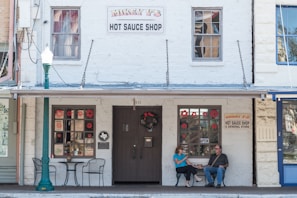 A quaint storefront with a sign reading 'Mikey V's Hot Sauce Shop' displays two windows adorned with red flower decals. Two people are seated in front of the shop at a small table, under a partially shaded awning. A green lamppost is positioned on the sidewalk, contributing to the vintage charm.