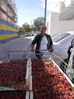 A person standing next to a cart filled with fresh strawberries, positioned outdoors near a construction site with a building under renovation. The cart has several crates stacked with strawberries, and a car is parked nearby. A bright blue and yellow tarp covers part of the building, and trees can be seen in the background with a clear sky overhead.
