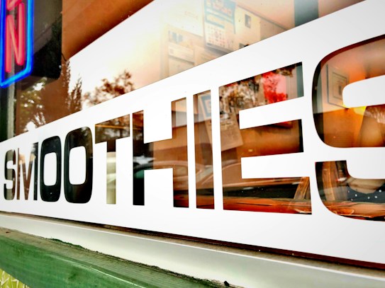 The image features a close-up view of a glass window with the word 'SMOOTHIES' prominently displayed in bold black and white letters. In the reflection on the glass, there is a glimpse of indoor decor and some blurred outdoor foliage, suggesting a pleasant environment.