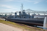 A large naval ship is docked beside a concrete embankment with a bridge in the background. The ship, painted in shades of grey, features various antennas and equipment on its deck, with a number 661 visible on the bow. The weather is clear with a blue sky, and the water beside the ship is calm.