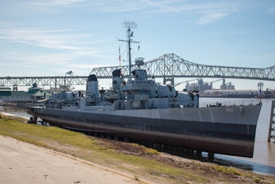 A large naval ship is docked beside a concrete embankment with a bridge in the background. The ship, painted in shades of grey, features various antennas and equipment on its deck, with a number 661 visible on the bow. The weather is clear with a blue sky, and the water beside the ship is calm.