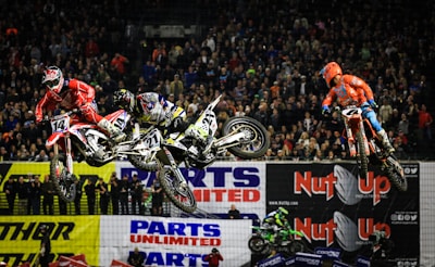 Three motocross riders are airborne in an indoor arena in front of a large crowd. The riders are dressed in colorful gear, including red and orange suits, as they perform jumps on dirt bikes. The background features banners with advertisements, and the spectators are watching the action closely.