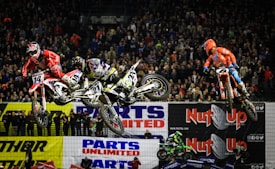 Three motocross riders are airborne in an indoor arena in front of a large crowd. The riders are dressed in colorful gear, including red and orange suits, as they perform jumps on dirt bikes. The background features banners with advertisements, and the spectators are watching the action closely.
