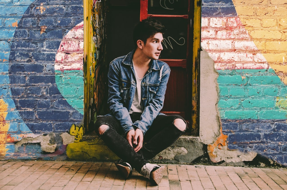 man in blue denim jacket and black pants sitting on doorway