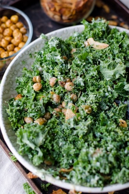 A close-up view of a salad primarily composed of fresh green kale, garnished with roasted chickpeas and possibly croutons or nuts. There are two small glass containers in the background filled with additional roasted chickpeas and what appear to be nuts or seeds.