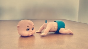 On a wooden surface, there is a toy figure with a detachable head next to its body. The head has large eyes and a mischievous expression. The body is positioned in a crawling stance, wearing blue shorts.
