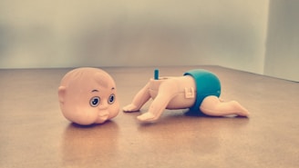 On a wooden surface, there is a toy figure with a detachable head next to its body. The head has large eyes and a mischievous expression. The body is positioned in a crawling stance, wearing blue shorts.