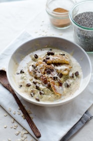 A bowl of creamy oatmeal topped with caramelized banana slices, chocolate chips, and a drizzle of sauce. Beside the bowl, there is a wooden spoon, some scattered oats, a jar of chia seeds, and a jar of cinnamon or another spice on a light-colored cloth.
