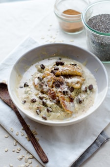 A bowl of creamy oatmeal topped with caramelized banana slices, chocolate chips, and a drizzle of sauce. Beside the bowl, there is a wooden spoon, some scattered oats, a jar of chia seeds, and a jar of cinnamon or another spice on a light-colored cloth.