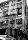 A multi-story residential building with balconies enclosed by metal grills. Laundry is hung outside on some balconies. Below the building are various makeshift tarpaulin-covered structures with posters and signs. An air conditioning unit is visible on one of the balconies.