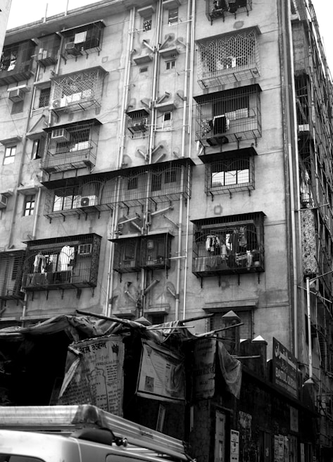 A multi-story residential building with balconies enclosed by metal grills. Laundry is hung outside on some balconies. Below the building are various makeshift tarpaulin-covered structures with posters and signs. An air conditioning unit is visible on one of the balconies.