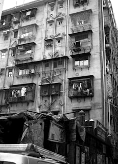 A multi-story residential building with balconies enclosed by metal grills. Laundry is hung outside on some balconies. Below the building are various makeshift tarpaulin-covered structures with posters and signs. An air conditioning unit is visible on one of the balconies.