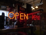 A neon 'OPEN' sign in bright orange is prominently displayed in the foreground. Behind it, there are various objects including a black Elvis poster, a framed picture on the wall, hanging light fixtures, and a collection of items that appear to be in a vintage or antique shop setting.