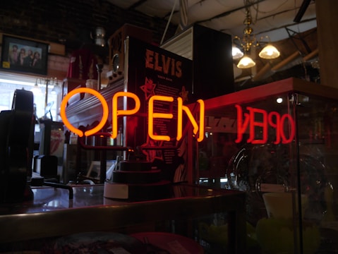 A neon 'OPEN' sign in bright orange is prominently displayed in the foreground. Behind it, there are various objects including a black Elvis poster, a framed picture on the wall, hanging light fixtures, and a collection of items that appear to be in a vintage or antique shop setting.