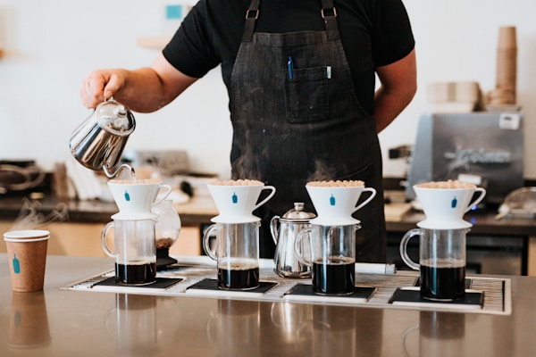 A barista in a black shirt and apron is making pour-over coffee using four glass carafes with white ceramic drippers. A stainless steel kettle is used to pour hot water over coffee grounds. There is a takeaway coffee cup on the counter and an espresso machine in the background.